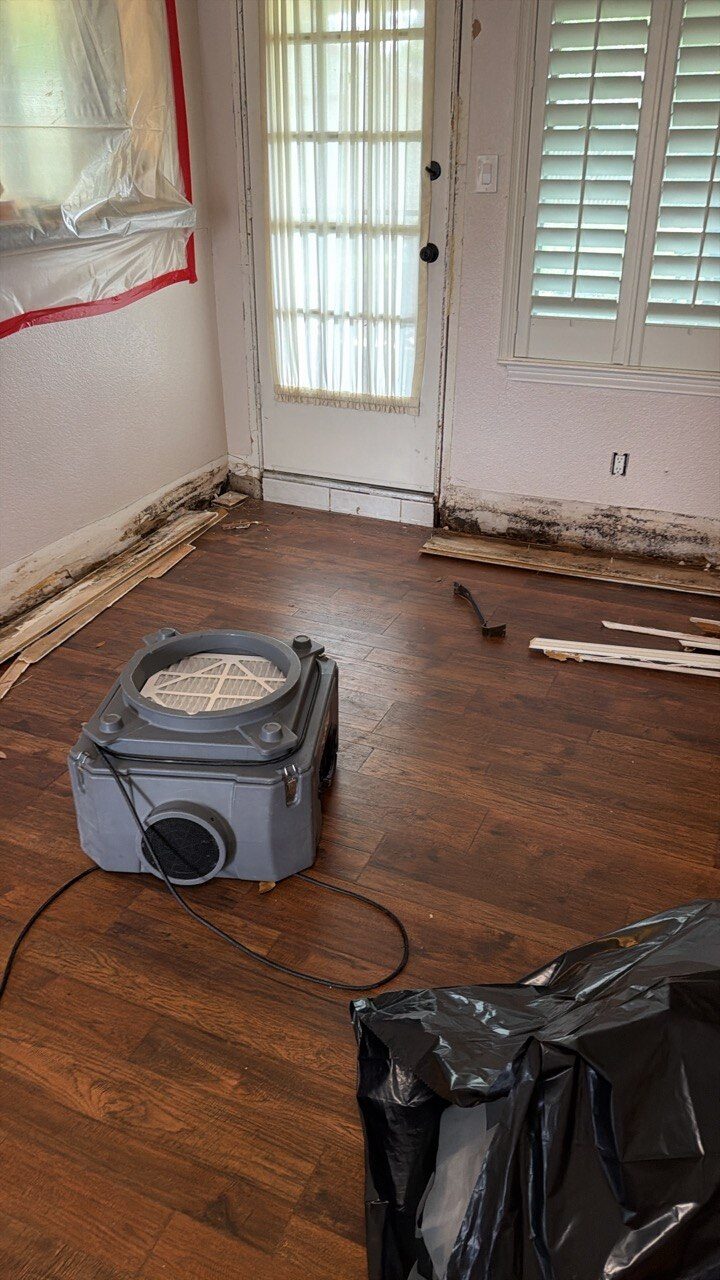 HEPA air scrubber running on the floor of a flood-cut room with an exterior glass door, drawing contaminated air through a HEPA filter to keep the work area safe