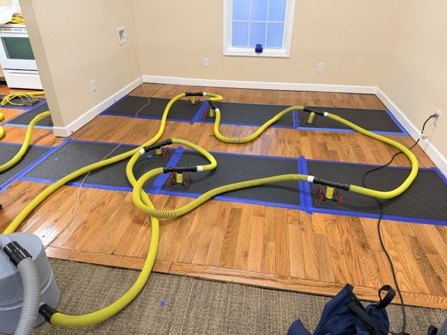 Hardwood flooring drying mats taped down across a kitchen floor with hoses running to extraction equipment after a hidden appliance leak.
