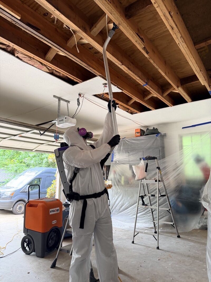 Flood Titan technician in full Tyvek PPE and P100 respirator performing Category 3 garage ceiling demolition with industrial dehumidifier and containment poly during DFW water damage restoration