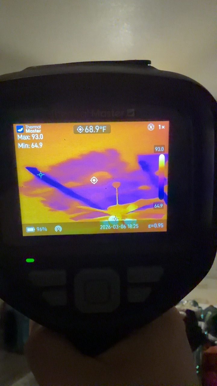 Thermal imaging camera held up to a textured ceiling showing a cooler purple bloom where hidden moisture has tracked along the drywall above a recessed light.